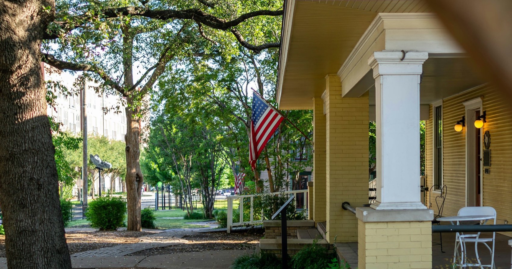 An American flag hanging from the porch of a house on a quiet, tree-lined street in warm afternoon light.
